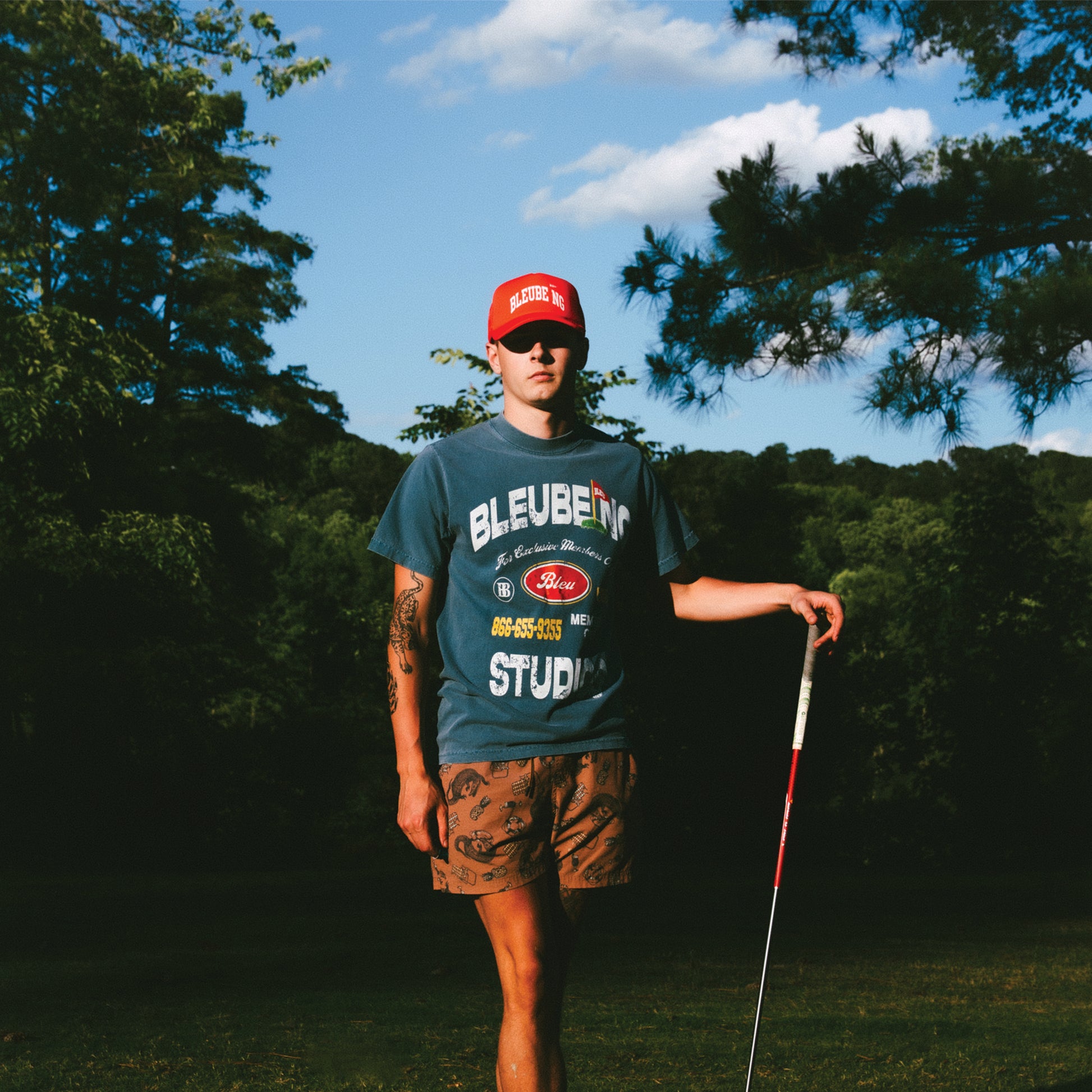 model holding golf club wearing slate blue bleubeing studios graphic t shirt with bold print on front and red mesh trucker hat with sky background.