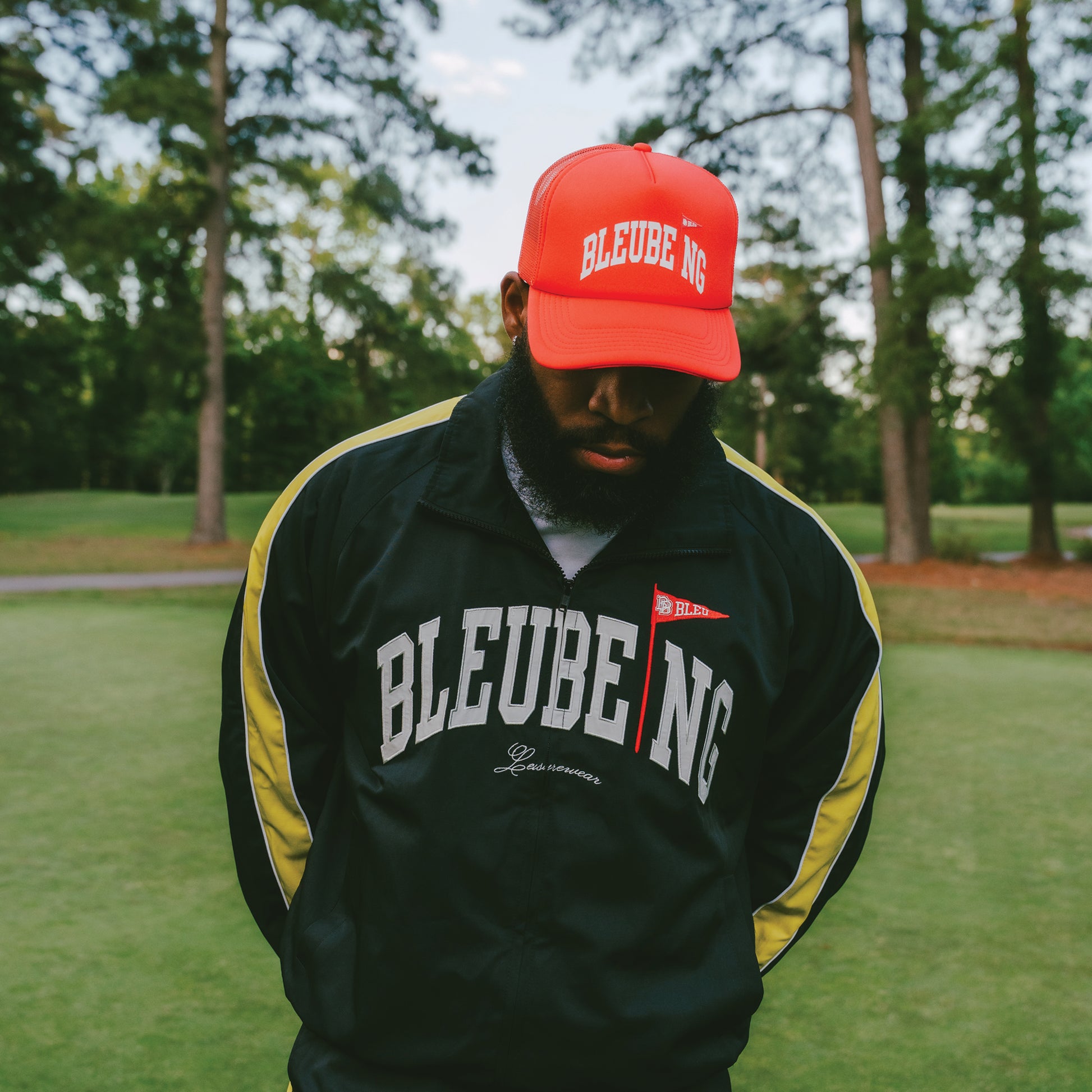 male wearing a athletic windbreaker jacket with a red trucker hat and sky and grass in background