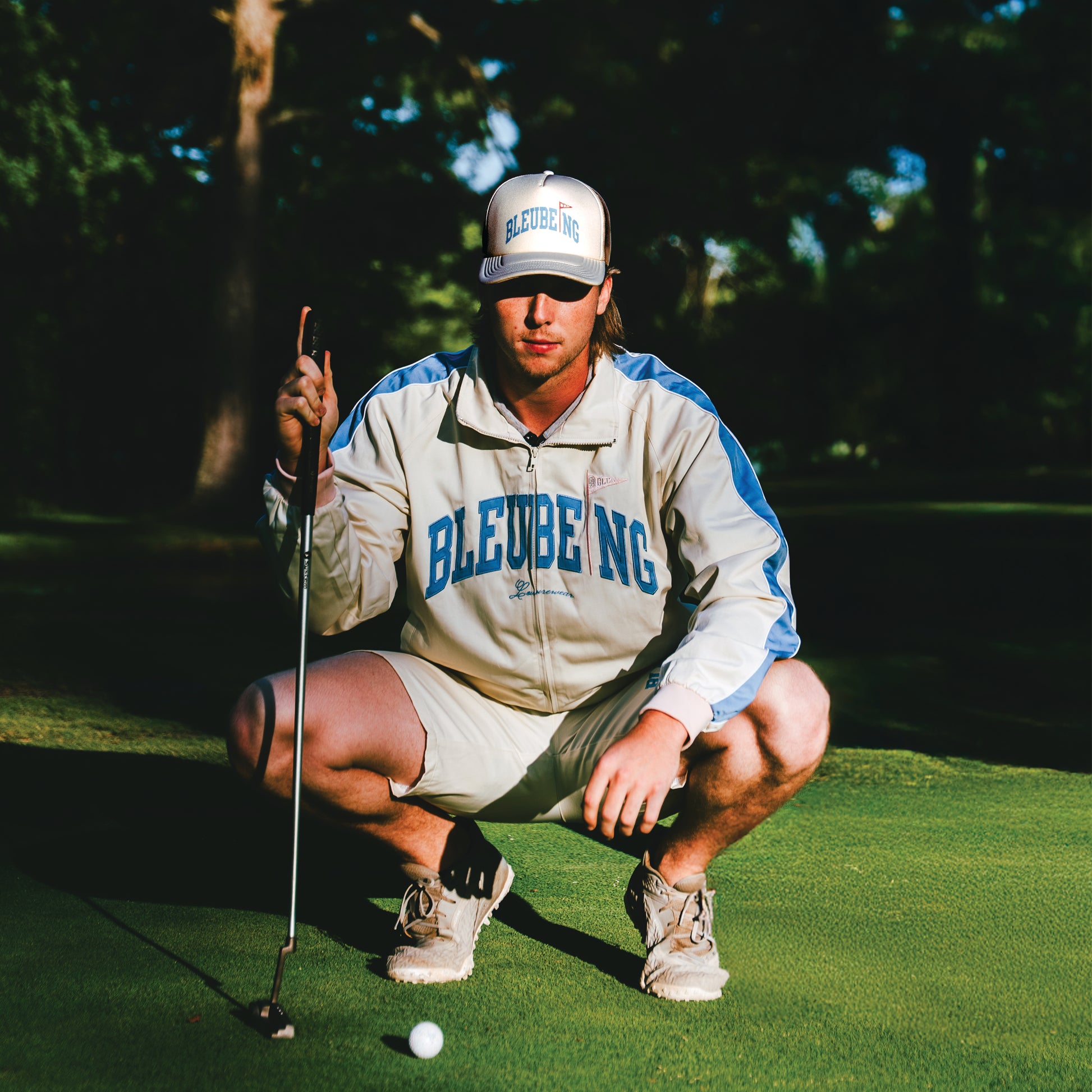 model at golf course holding golf club wearing a cream athletic windbreaker set shorts and jacket with cream mesh trucker hat.