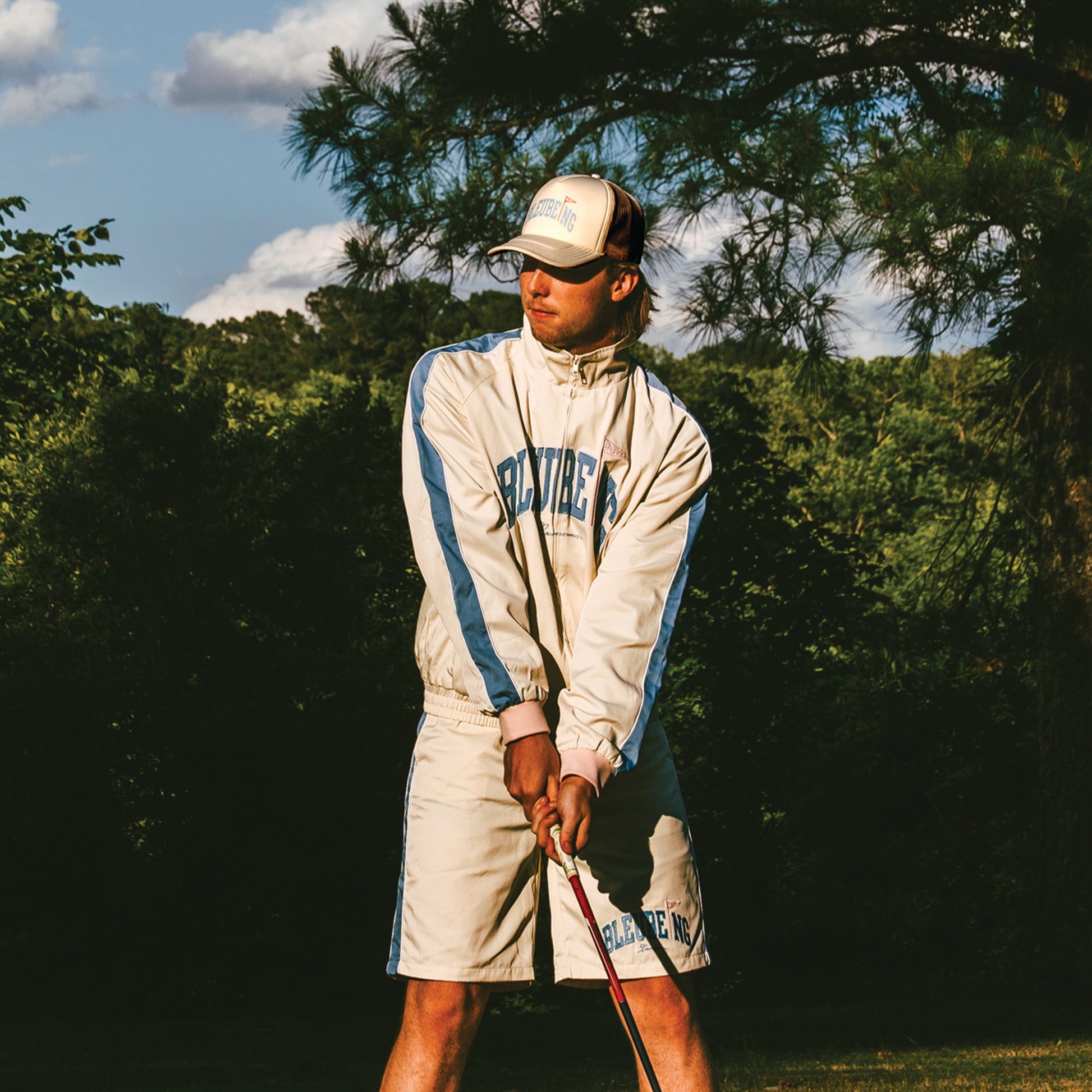 model at golf course holding golf club wearing a cream athletic windbreaker set shorts and jacket with cream mesh trucker hat.