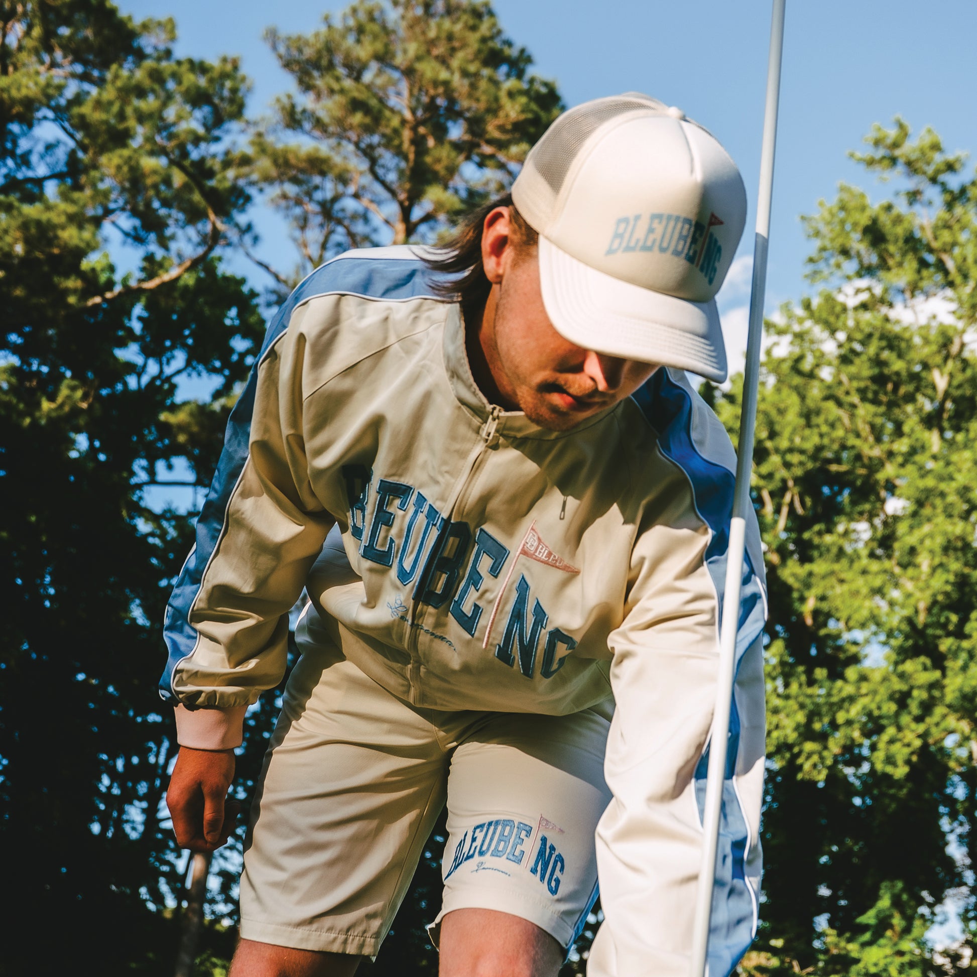 model at golf course wearing a cream athletic windbreaker set shorts and jacket with cream mesh trucker hat.